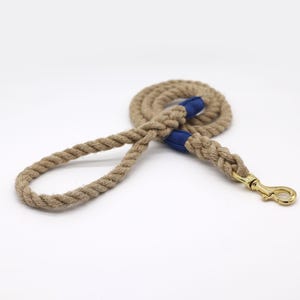 May include: A tan rope dog leash with blue fabric accents and a gold-colored metal clasp. The leash is coiled and lies on a white surface. The rope is thick and appears durable.