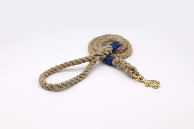 May include: A tan rope dog leash with blue fabric accents and a gold-colored metal clasp. The leash is coiled and lies on a white surface. The rope is thick and appears durable.