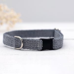 May include: A gray cat collar with a woven texture. The collar features a black plastic buckle and a silver-colored metal D-ring. The collar is designed for pets.
