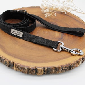 May include: Black dog leash with a silver metal clasp and handle. The leash is made of a woven material and features a small rectangular tag with the text "JUST HEMP - BY WIGGLYWOODS". The leash is displayed on a wooden surface.