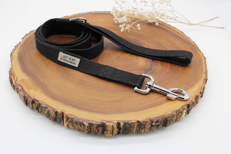 May include: Black dog leash with a silver metal clasp and handle. The leash is made of a woven material and features a small rectangular tag with the text "JUST HEMP - BY WIGGLYWOODS". The leash is displayed on a wooden surface.