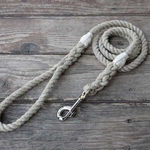 May include: A beige rope dog leash with a silver-colored metal clasp. The leash is made of thick, braided rope and features white fabric accents at the handle and clasp attachment points. The leash is coiled on a weathered wooden surface.