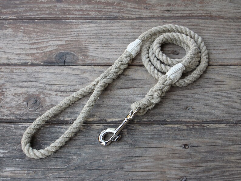 May include: A beige rope dog leash with a silver-colored metal clasp. The leash is made of thick, braided rope and features white fabric accents at the handle and clasp attachment points. The leash is coiled on a weathered wooden surface.