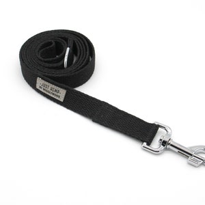 May include: A black dog leash made of woven material with a silver metal clasp. The leash is rolled up and has a small rectangular tag that reads "- JUST HEMP - BY WIGGLYWOODS."