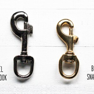 May include: Two snap hooks, one nickel and one brass, displayed side-by-side. The nickel snap hook is silver-colored, while the brass snap hook has a gold finish. Text below each hook identifies them as "NICKEL SNAP HOOK" and "BRASS SNAP HOOK."