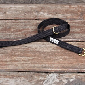 May include: A black dog leash with a brass-colored swivel clasp and a matching brass-colored ring. The leash is made of a woven material and has a small white tag with black text.