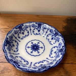 May include: A small, white and blue porcelain dish with a scalloped edge. The dish features a central floral design and intricate blue patterns around the rim. The dish is placed on a wooden surface.