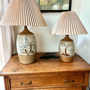 May include: Two matching ceramic table lamps with beige pleated shades. The lamp bases are a speckled beige with blue floral designs. The lamps are placed on a wooden table with drawers.