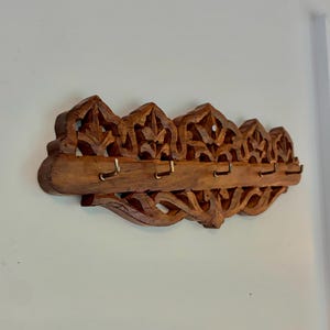 May include: A wall-mounted wooden coat rack featuring intricate hand-carved floral patterns along the top and bottom edges. The horizontal brown wood piece includes five small, evenly spaced brass-colored metal hooks for hanging items.