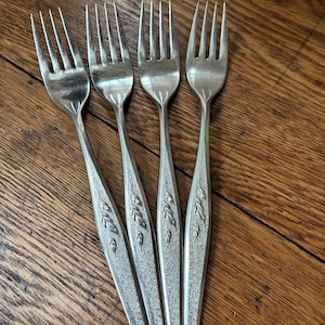 May include: A set of four silver-toned metal forks featuring a textured handle design with an embossed leaf pattern. Each fork has four tines and a tapered handle that widens toward the center before narrowing at the base, showcasing a metallic finish.