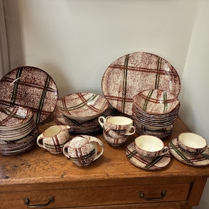 May include: A collection of vintage dishware with a red, green, and white plaid pattern. The set includes plates, bowls, cups, and saucers. The dishware is arranged on a wooden surface.