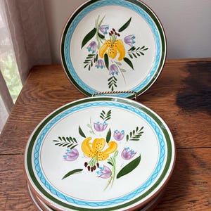 May include: Set of decorative ceramic plates featuring a floral design. Each plate has a white center with a yellow lily and purple flowers. The rims have a green and blue border, and a brown edge.