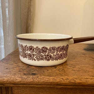 May include: White enamel saucepan with a brown floral pattern and a brown handle. The saucepan is sitting on a wooden surface.