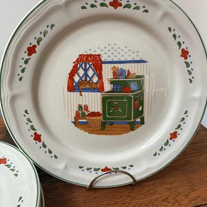 May include: A round white ceramic dinner plate with a green rim and a decorative border of red and green floral motifs. The center features a kitchen scene with a green stove, a window with red curtains, a cat, and jars of preserved food.