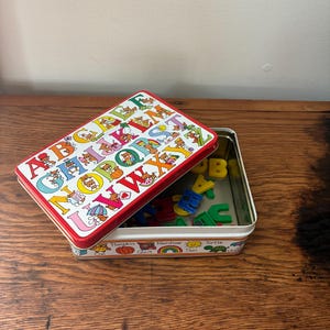 May include: A vintage alphabet learning set in a rectangular tin. The tin has a red rim and a white background with colorful illustrations of letters and bears. The lid is open, revealing colorful plastic letters inside.