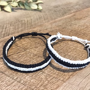 May include: Two friendship bracelets, one black and white and one white and black, are woven together with a simple knot pattern. The bracelets are lying on a wooden surface.