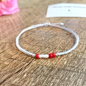 May include: A silver beaded bracelet with red and white beads. The bracelet is tied with a silver cord.