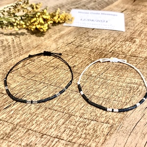 Puede incluir: Dos pulseras de cuentas, una negra y otra blanca, con detalles plateados. Las pulseras están sobre una superficie de madera.