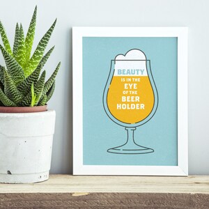 May include: A framed print with a light blue background features a beer glass illustration filled with yellow liquid and white foam. The text "BEAUTY IS IN THE EYE OF THE BEER HOLDER" is printed in the glass. A potted succulent plant sits to the left.
