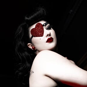 May include: A black lingerie set featuring a high-waisted sheer brief with garter straps and a matching bra. The ensemble is accented by a red heart-shaped eye patch covered in small sparkling crystals, red stud earrings, and bold red lipstick.