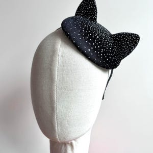 May include: A black cat ear headband with silver glitter accents. The ears are upright and attached to a thin black headband. The accessory is displayed on a mannequin head.