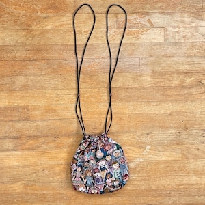 Puede incluir: Una pequeña bolsa con cordón y un colorido estampado de muñecas. La bolsa tiene un cordón negro para llevarla o colgarla. El fondo es una superficie de madera de color marrón claro.