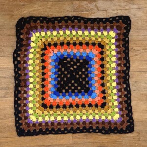 Op de afbeelding: Een vierkante gehaakte deken met een veelkleurig granny square patroon. Het ontwerp bestaat uit concentrische ringen van zwart, bruin, paars, geel, limoengroen, oranje en blauw garen, afgewerkt met een brede zwarte geschulpte rand.