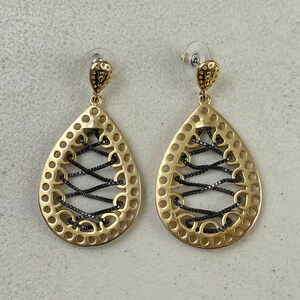 May include: A pair of gold-tone earrings with a teardrop shape. The earrings feature a black criss-cross design.