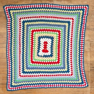 Op de afbeelding: Een vierkante gehaakte deken met een herhalend granny square patroon met concentrische randen in afwisselende kleuren, waaronder donkerblauw, rood, lichtgroen, geel, lichtblauw en wit, wat zorgt voor een levendig en geometrisch textielontwerp.