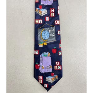 May include: Navy blue tie with a school theme. The tie features illustrations of books, apples, backpacks, and a globe with the equation 17-7=10. The tie also has the letters A, B, C, D, E, F, G, Y, and Z.