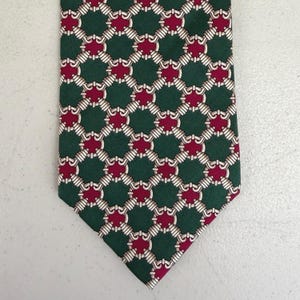 May include: A green necktie with a repeating pattern of burgundy and white interlocking shapes. The tie is made of a smooth fabric and has a classic pointed end. The pattern is symmetrical and covers the entire surface of the tie.