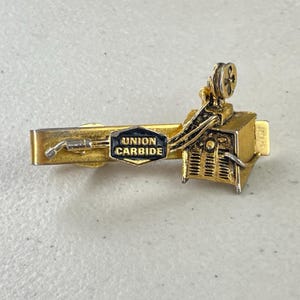 May include: Gold-tone tie clip featuring a stylized welding machine with a black and gold 'UNION CARBIDE' logo.