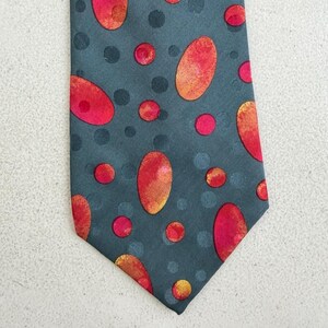 May include: A necktie featuring a dark gray background with a pattern of scattered, solid dark gray circles and larger, vibrant red and orange oval shapes. The design is distributed across the entire surface of the pointed fabric accessory.