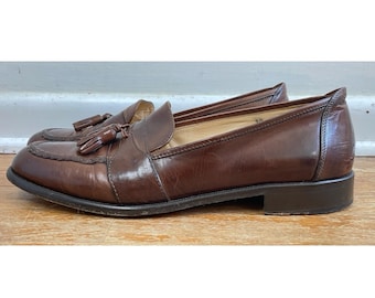 Pode incluir: Um par de mocassins de couro castanho com detalhes de borlas. Os sapatos têm um design clássico com bico redondo e salto baixo. O couro tem um acabamento brilhante e os sapatos estão sobre uma superfície de madeira.