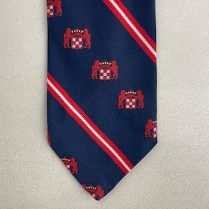 May include: Navy blue tie with a repeating pattern of red crests and diagonal red and white stripes. The tie is made of a smooth fabric and has a classic pointed end. The crests feature a crown and two lions.
