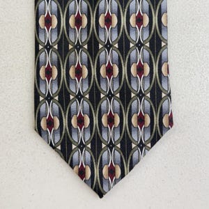 May include: A patterned necktie featuring a repeating oval design in black, gray, red, and tan. The tie has a pointed end and is likely made of silk or a similar fabric. The overall pattern is symmetrical and geometric.