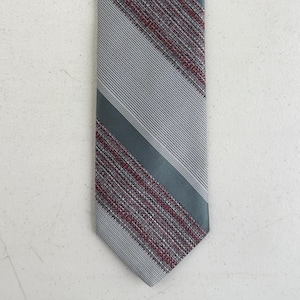 May include: A necktie featuring a diagonal striped pattern with alternating sections of light gray fine-ribbed fabric and a textured weave of dark gray and deep red threads. The design consists of clean lines and a pointed bottom edge.