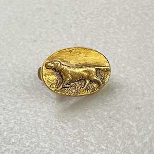 May include: A gold-toned oval pin featuring a detailed relief of a dog running through tall grass under a sky with birds. The pin has a classic, vintage aesthetic, suitable for collectors or those who appreciate animal-themed jewelry.