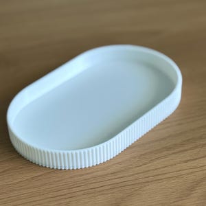 Può includere: Un vassoio ovale bianco con una base interna liscia e una parete esterna a coste verticali. Il vassoio presenta una finitura in tinta unita e un bordo rialzato, offrendo una superficie semplice e funzionale per piccoli oggetti.