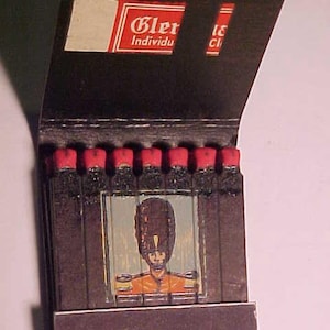 May include: A vintage matchbook cover with a red and white label that reads "Glen Individual", featuring a black and white illustration of a person in a military uniform with a red hat. The matchbook has a black background and a red stripe along the bottom.