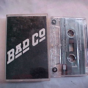 May include: A black cassette tape with a clear plastic case. The tape has the text "BAD CO" printed in white on the front.