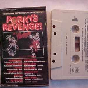 May include: A black cassette tape with a white label. The label has the text "PORKY'S REVENGE!" and the text "THE ORIGINAL MOTION PICTURE SOUNDTRACK" in white letters on a black background. The cassette tape is in a plastic case.