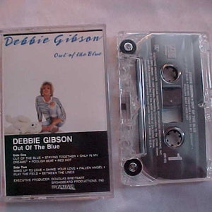 May include: A cassette tape case with a photo of Debbie Gibson on the front. The case is clear plastic and the photo is of Debbie Gibson in a white shirt and blue jeans. The text on the case reads "Debbie Gibson Out of the Blue".