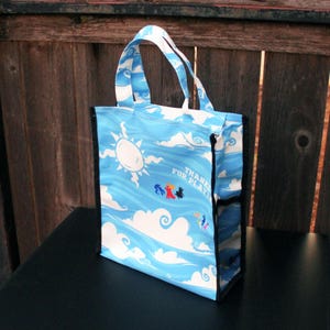 May include: A blue tote bag with a whimsical sky design featuring a sun, clouds, and waves. The bag has a black mesh side pocket and white handles. The text "THANKS FOR PLAYING" is visible on the front.