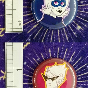 Puede incluir: Dos pines de bot&oacute;n de 2,5 cm que representan personajes de dibujos animados con cabello blanco. El primer pin tiene un fondo azul y presenta un personaje con una m&aacute;scara morada. El segundo pin tiene un fondo rojo y presenta un personaje con gafas de sol.