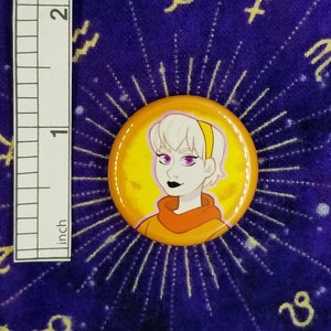 Puede incluir: Un bot&oacute;n amarillo y naranja con una imagen de dibujos animados de una persona con cabello blanco, ojos morados y una diadema amarilla. La persona lleva una camisa naranja.