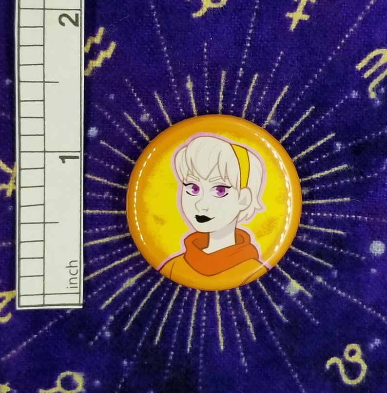 Puede incluir: Un bot&oacute;n amarillo y naranja con una imagen de dibujos animados de una persona con cabello blanco, ojos morados y una diadema amarilla. La persona lleva una camisa naranja.