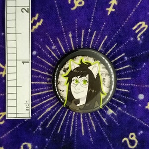 Puede incluir: Un bot&oacute;n blanco y negro con una imagen de dibujos animados de una persona con ojos verdes y cabello negro que lleva gafas. La persona tiene orejas de gato y lleva una camisa negra. El bot&oacute;n est&aacute; sobre un fondo morado con estrellas blancas.