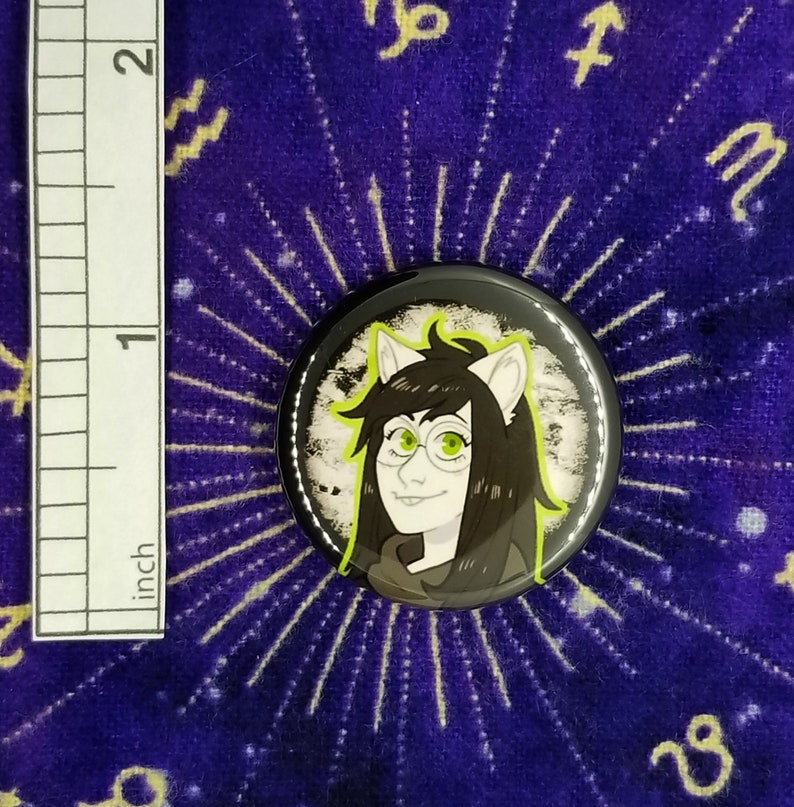 Puede incluir: Un bot&oacute;n blanco y negro con una imagen de dibujos animados de una persona con ojos verdes y cabello negro que lleva gafas. La persona tiene orejas de gato y lleva una camisa negra. El bot&oacute;n est&aacute; sobre un fondo morado con estrellas blancas.