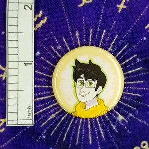 Puede incluir: Un bot&oacute;n amarillo con una ilustraci&oacute;n de dibujos animados de una persona con ojos verdes y cabello casta&ntilde;o que lleva una sudadera con capucha amarilla. La persona est&aacute; sonriendo.